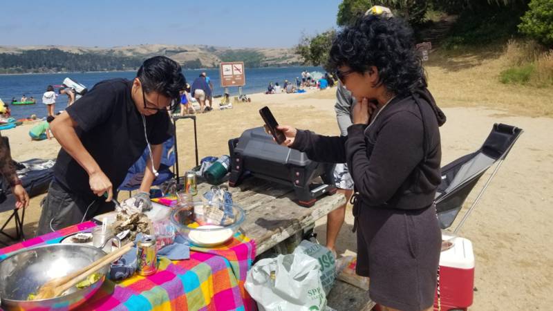 A woman holds up her cellphone to film a young man shucking oysters on the beach.