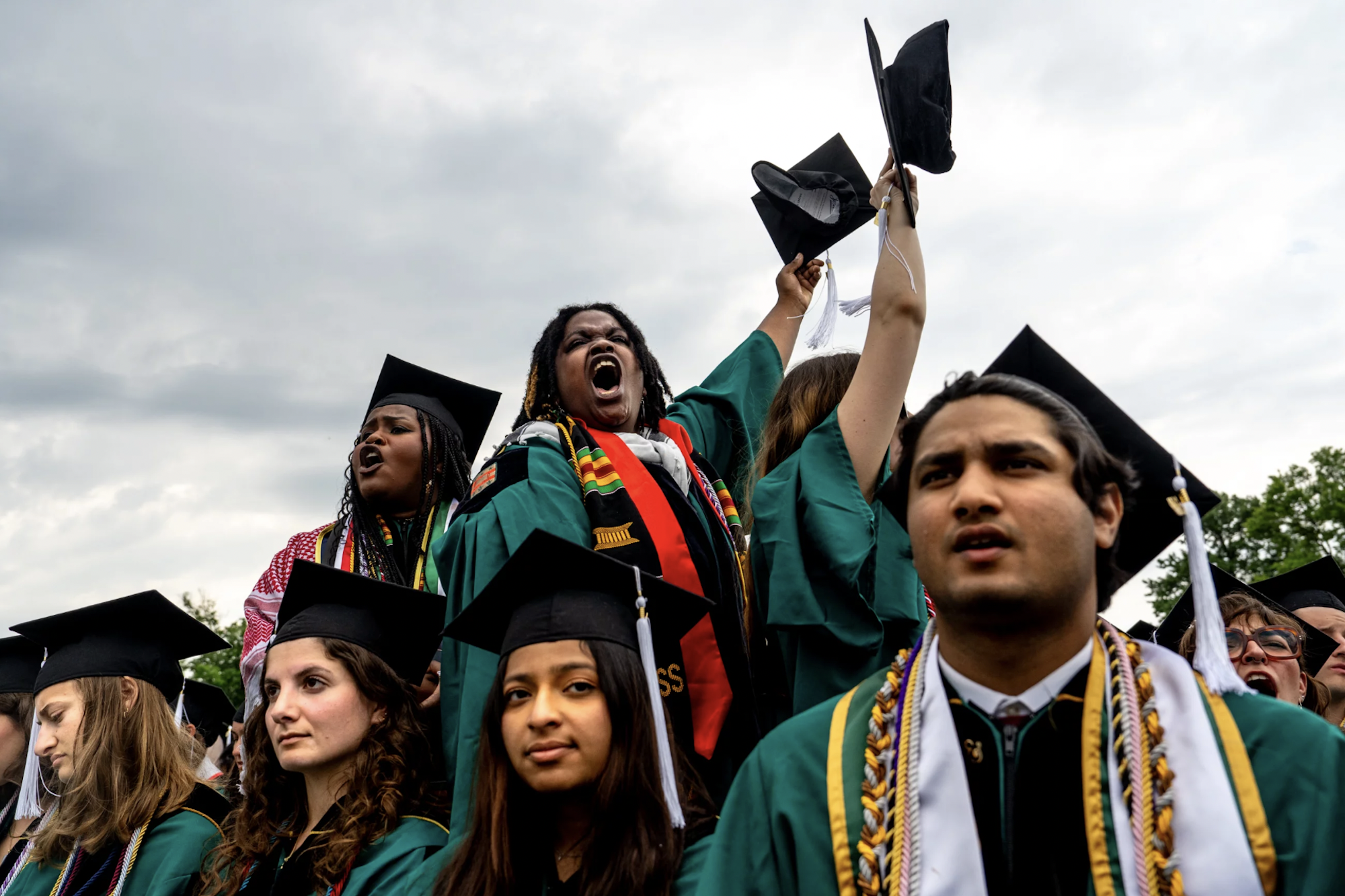 A diverse group of students wearing graduation caps and gowns, some stone faced, some shouting, stand close together.