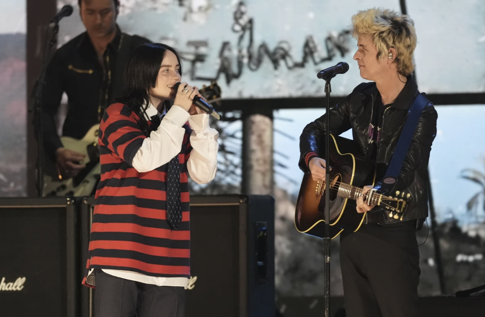 A young white woman with long black hair, wearing a red and black striped shirt sings into a microphone on stage, next to a white man with bleached hair wearing all black and playing a guitar.