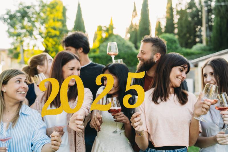 A group of 20-somethings stand in a garden smiling and drinking wine. Three of them are holding up numbers that spell out 2025.