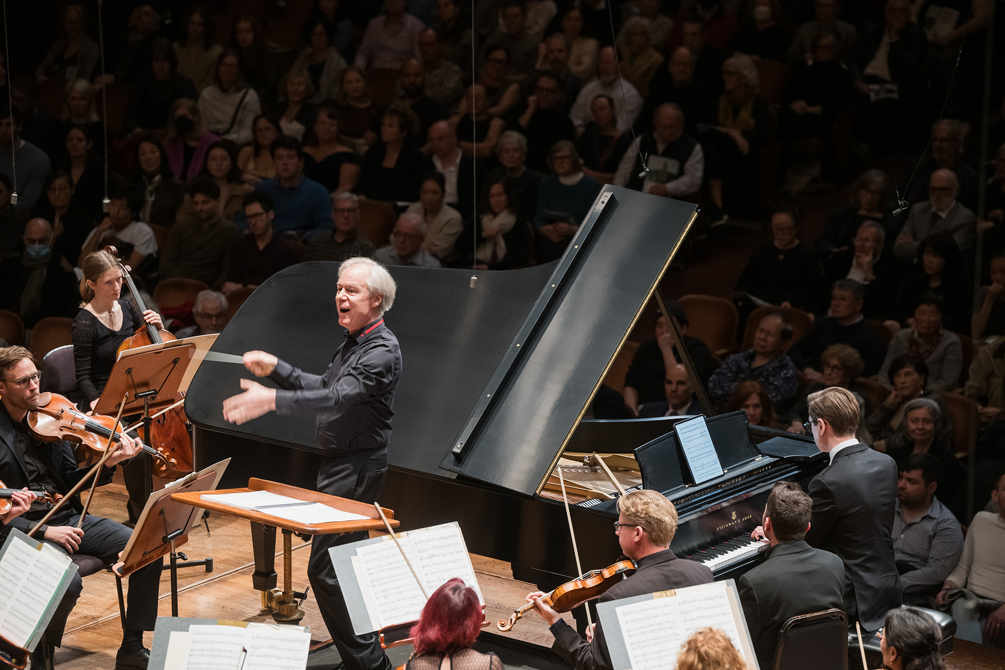 A man in a black shirt gesturing to an orchestra next to a piano and violins, with a seated audience in the background