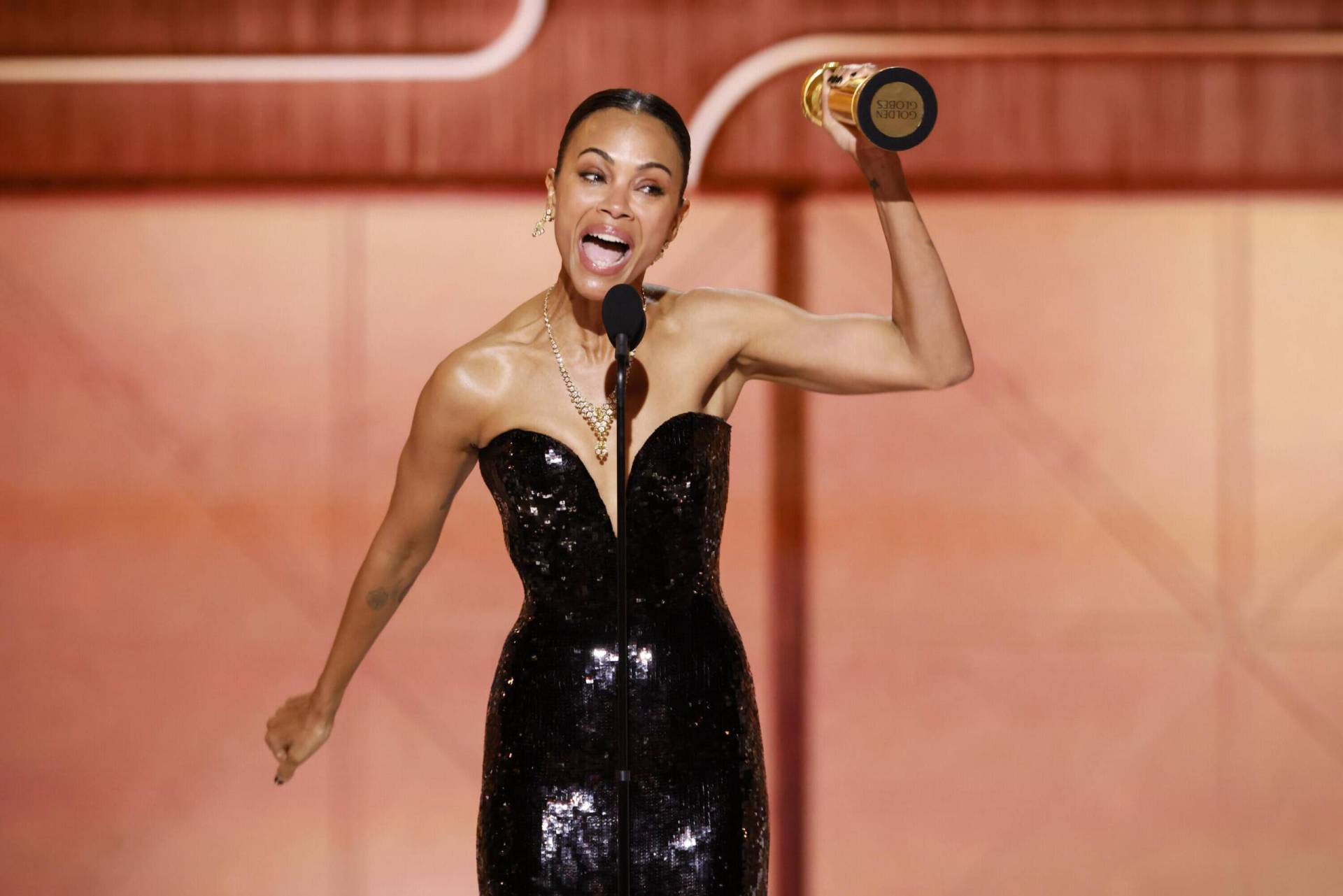 A very slender Black woman wearing a strapless black gown speaks into a microphone on stage, waving an award near her head.
