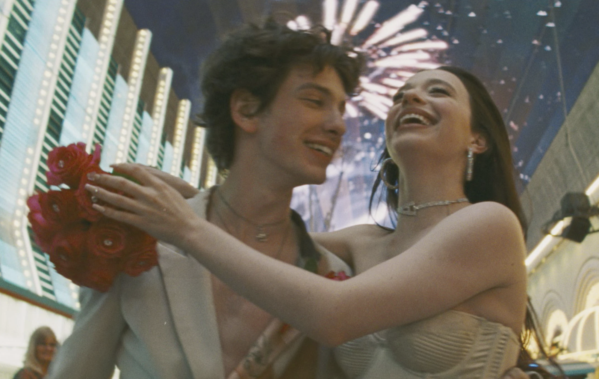 A young man and woman laugh and embrace against a backdrop of bright lights and fireworks.