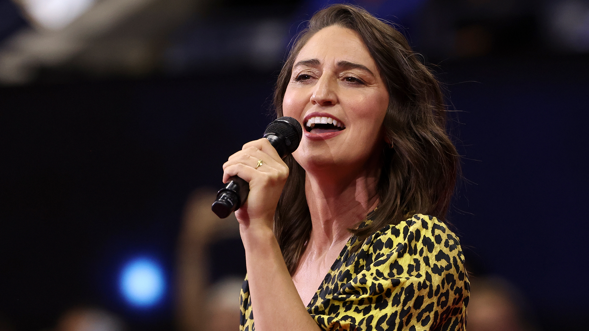 A brunette woman in a yellow and black spotted top sings into a microphone, looking to the left