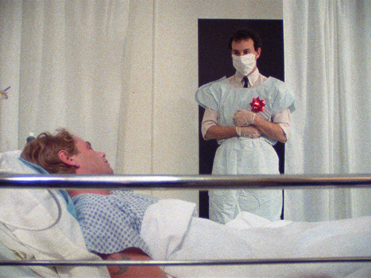 man in hospital smock and mask looks down at man on hospital bed
