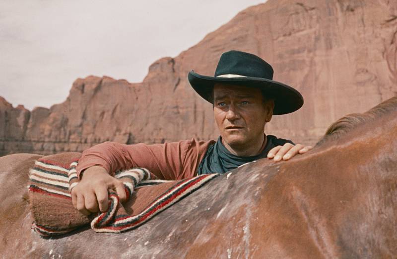 A man in a cowboy hat stands astride a horse against the red rock of the American desert