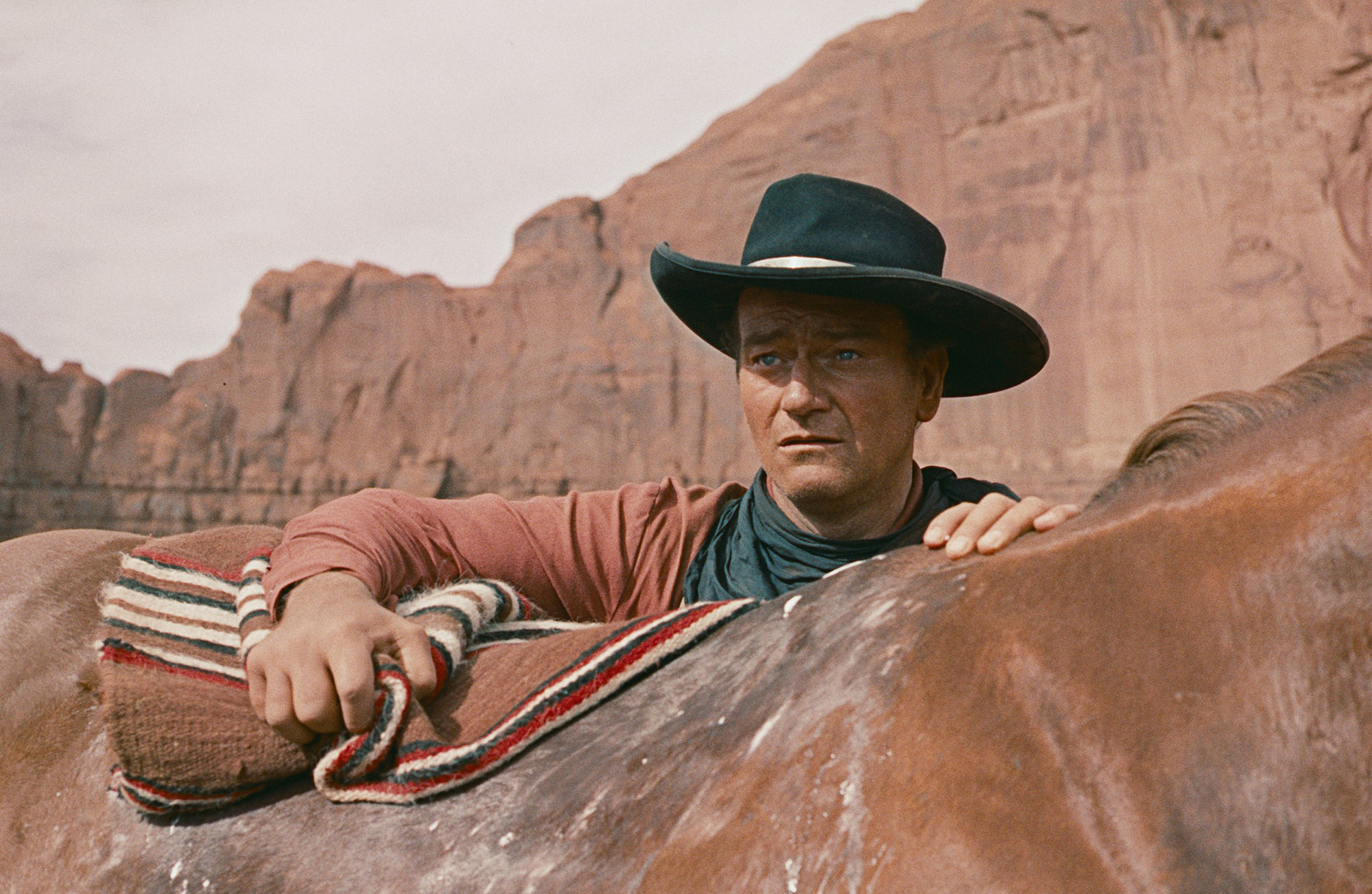 A man in a cowboy hat stands astride a horse against the red rock of the American desert
