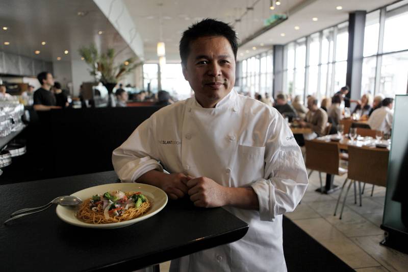 man in chef jacket smiles with noodle dish displayed at elbow in large restaurant