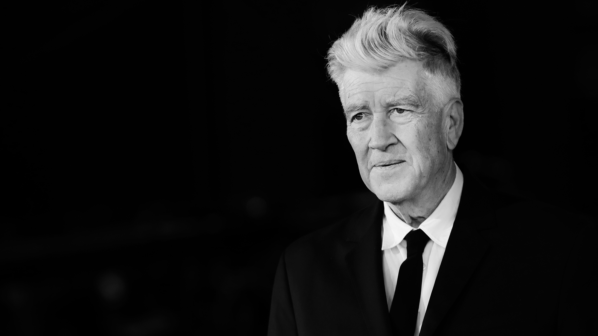 A man with grey hair in a suit and tie against a black backgroound
