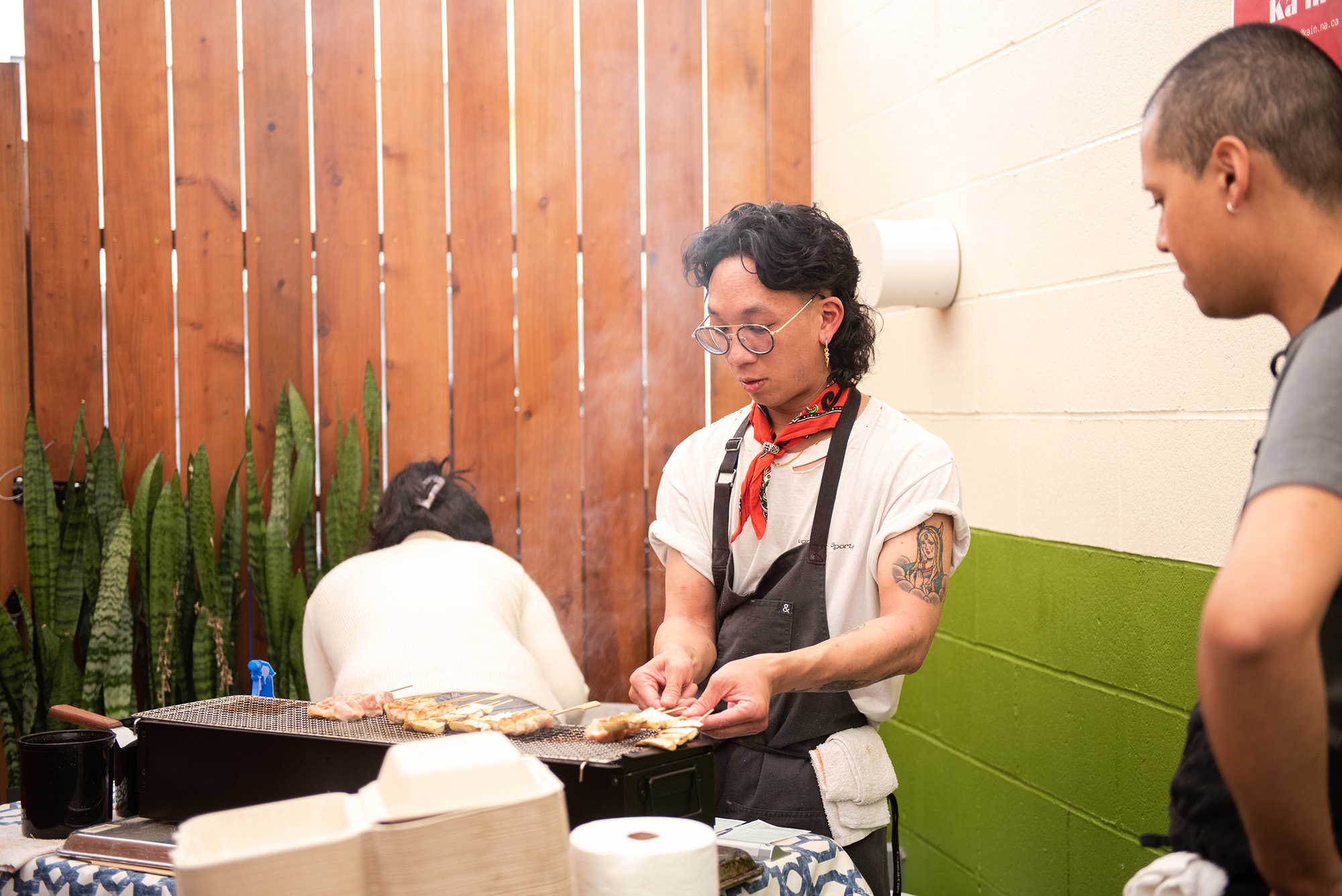 A tattooed street vendor cooks skewers over a portable grill.