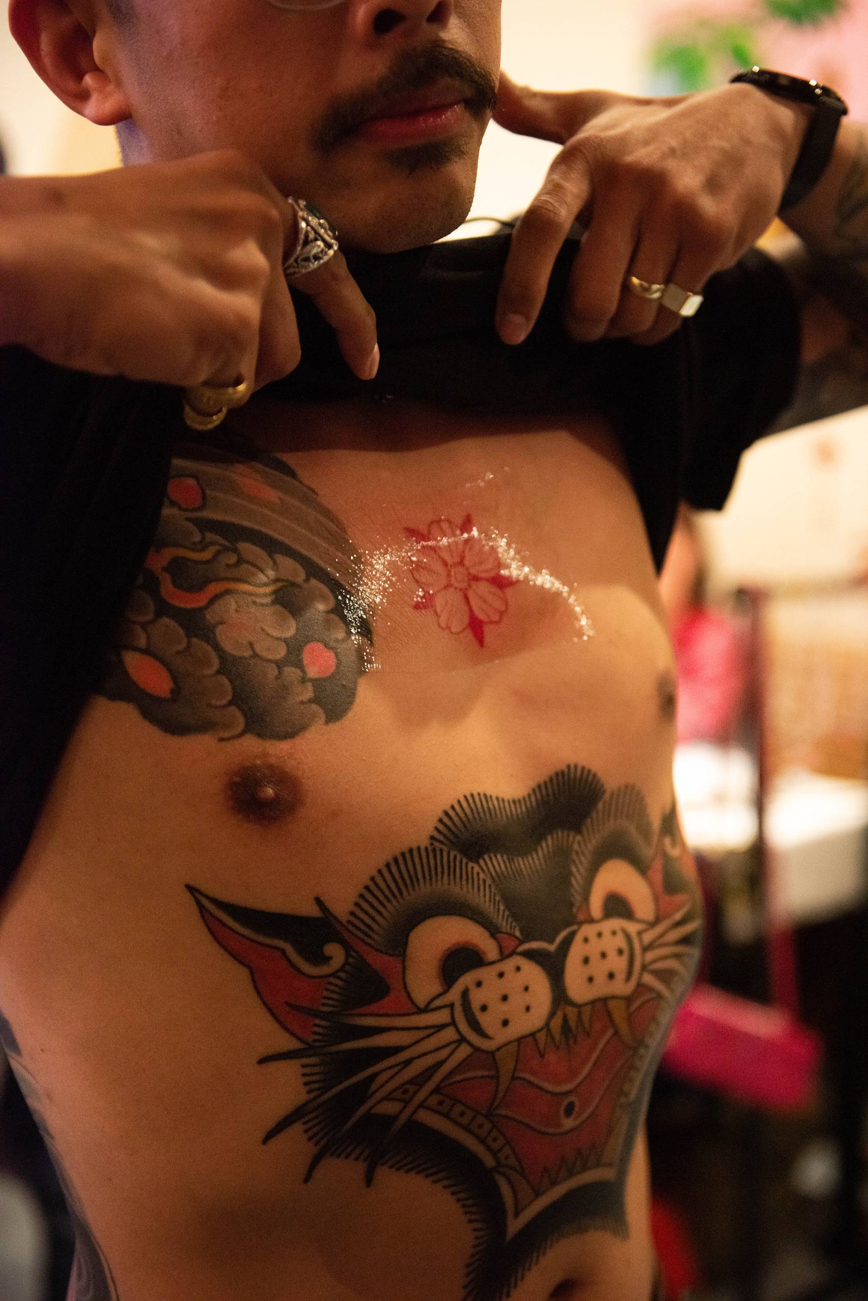 A man lifts up his shirt to reveal several tattoos, including a red flower on his sternum and a large cat across his abdomen. 