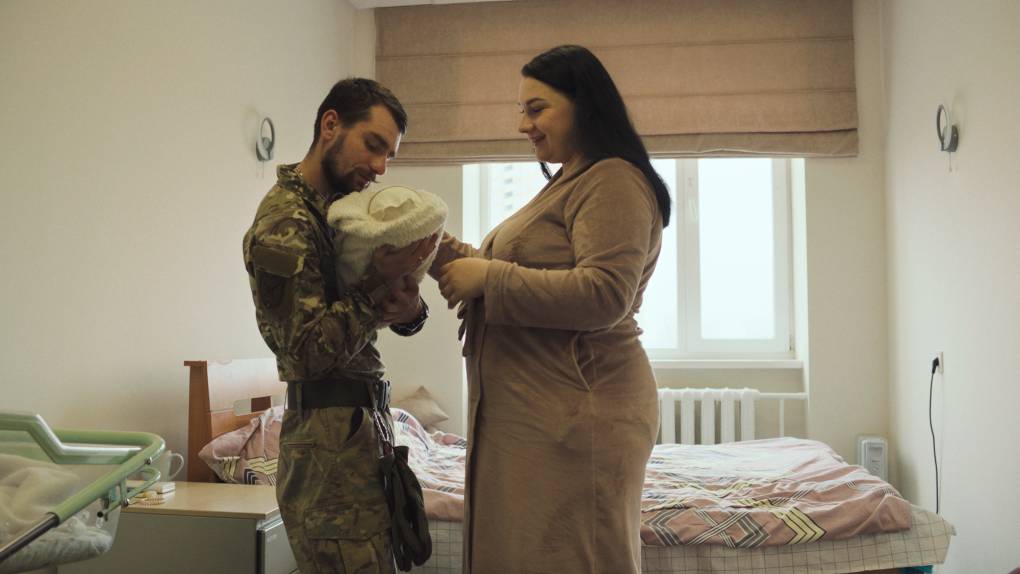 man in military uniform holds bundled baby, woman looks on smiling