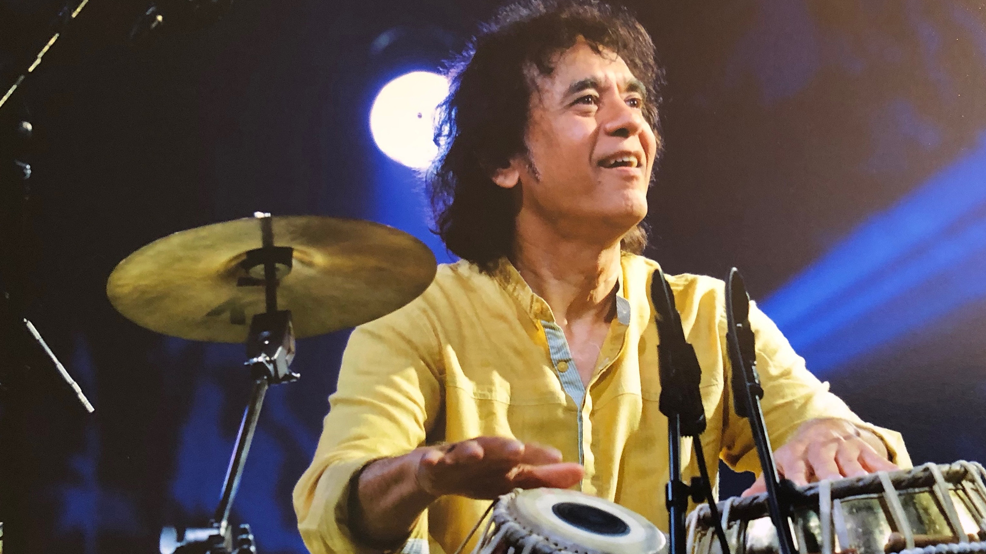 A man in a yellow top looks upward while playing round Indian drums