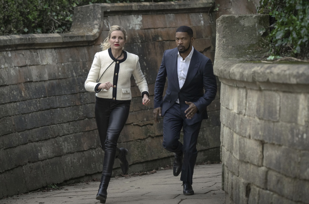 A white blonde woman wearing leather pants and a white Chanel jacket runs down a narrow path with a handsome Black man in a suit. They are surrounded by high stone walls.