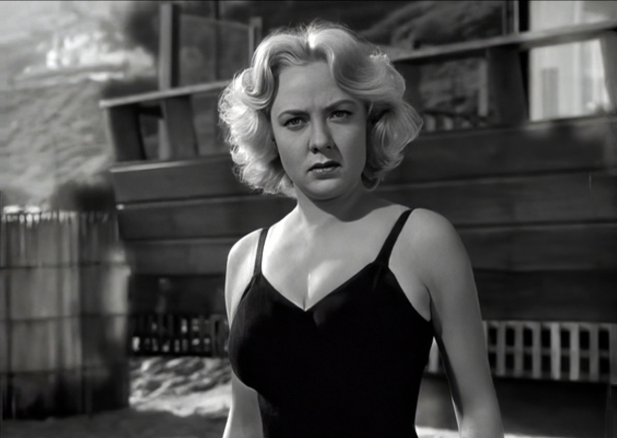 A woman stands outside in the sunlight, wearing a black bathing suit. She looks angry and frustrated.
