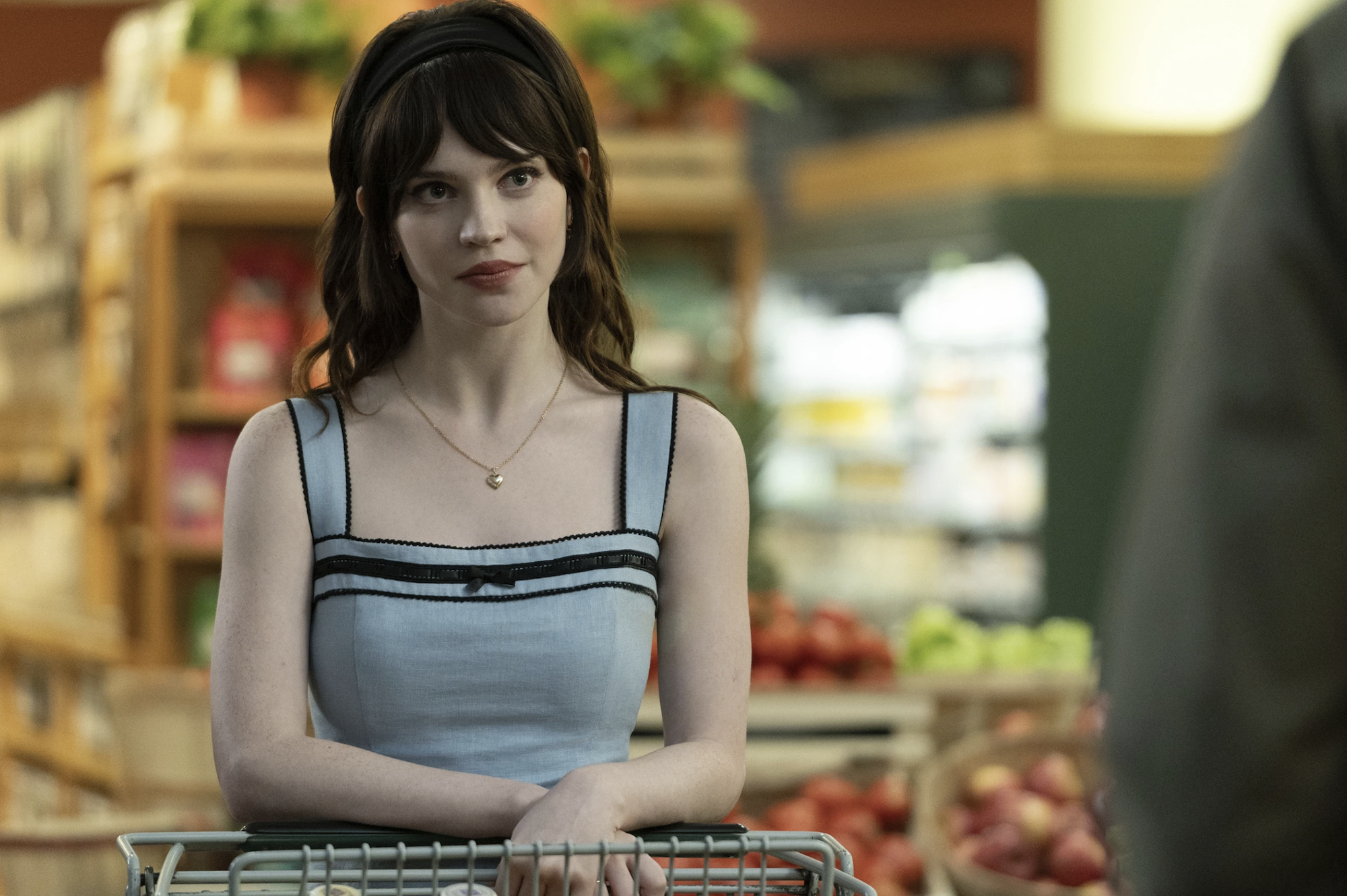 A beautiful pale young woman with cascading dark brown hair stands in a supermarket produce section behind a shopping cart. She is wearing a 1950s-style blue dress.