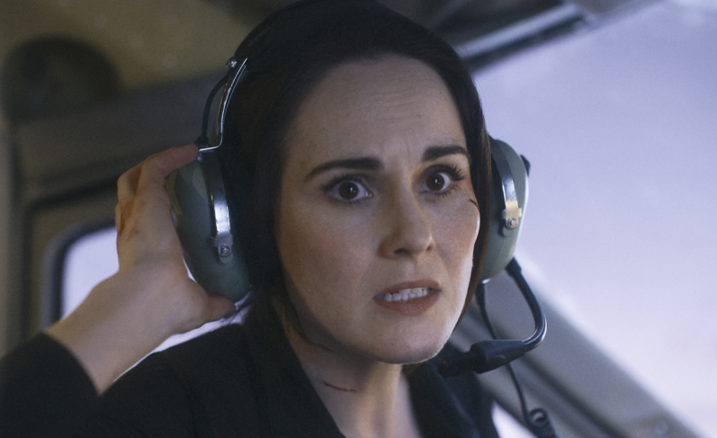 A white woman with black hair sits in a cockpit, wearing headphones and a concerned expression.