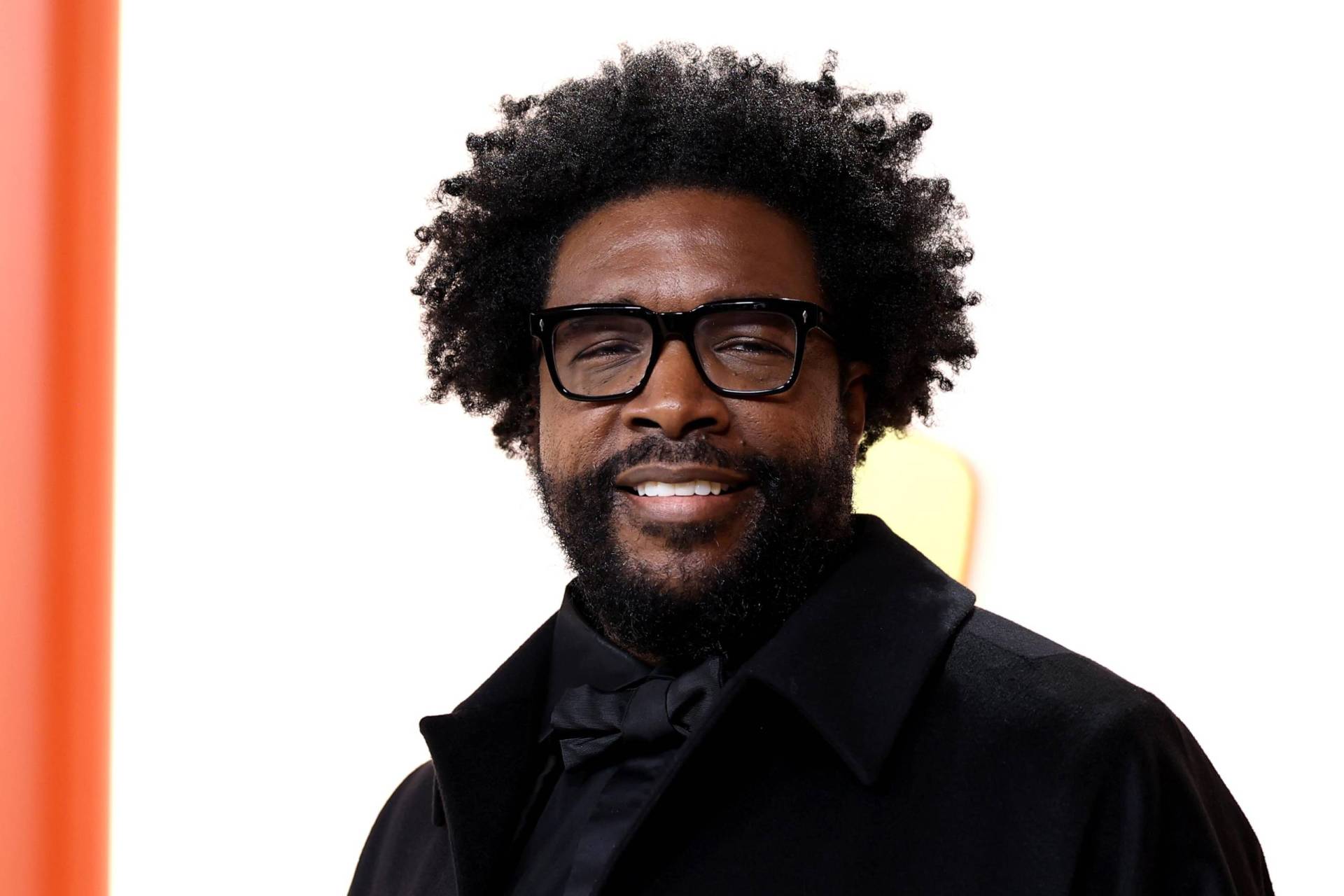 A Black man with need beard, black-rimmed glasses and mid-length afro smiles on a red carpet.