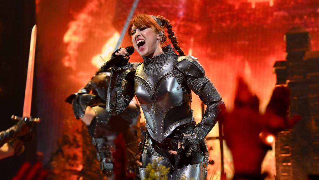 A woman with long braided red hair wearing a suit of armor screams into a microphone on stage.