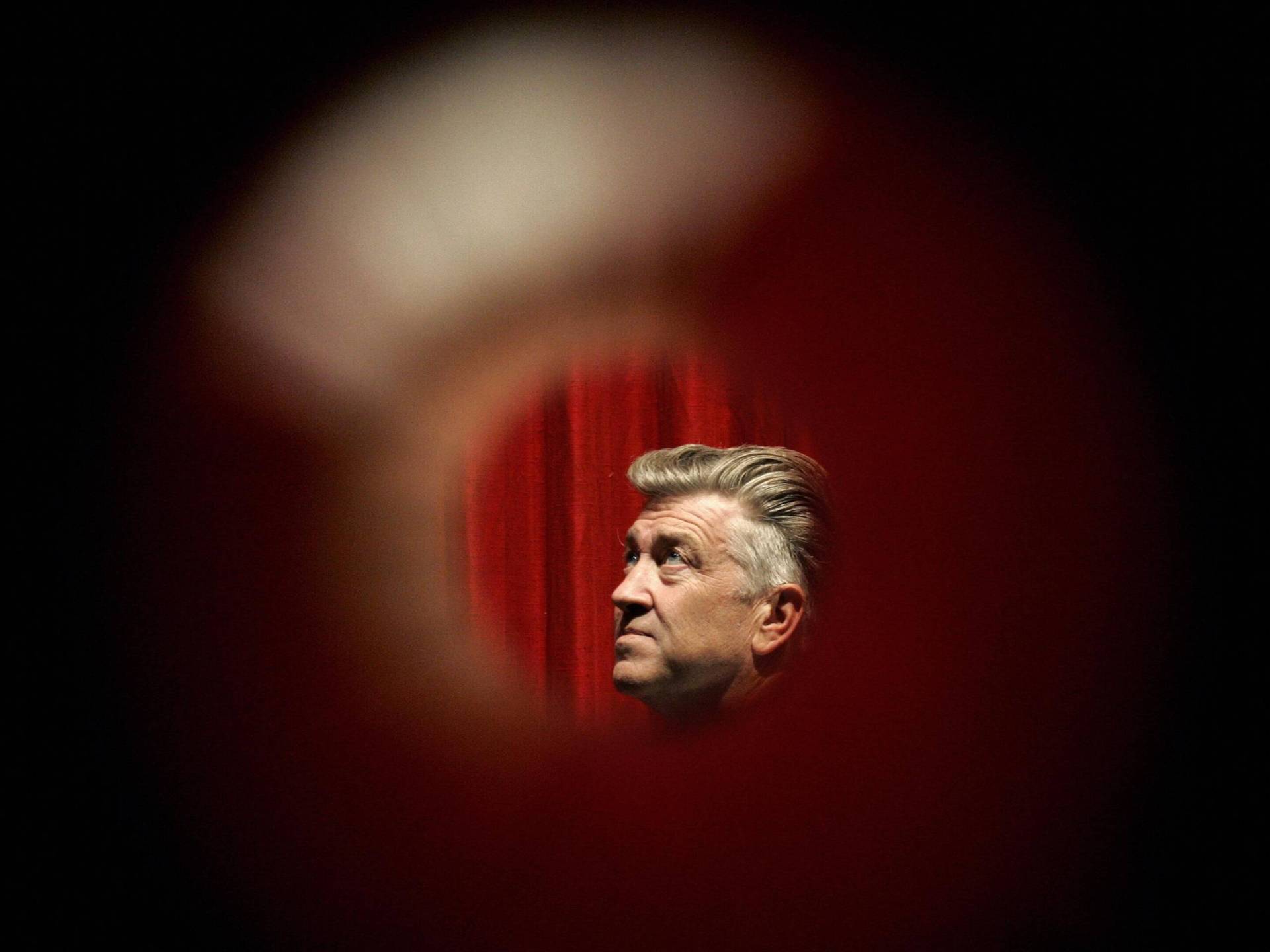 A man with grey hair is seen in side profile in front of a red velvet curtain. He is viewed through a circular peephole or lens.