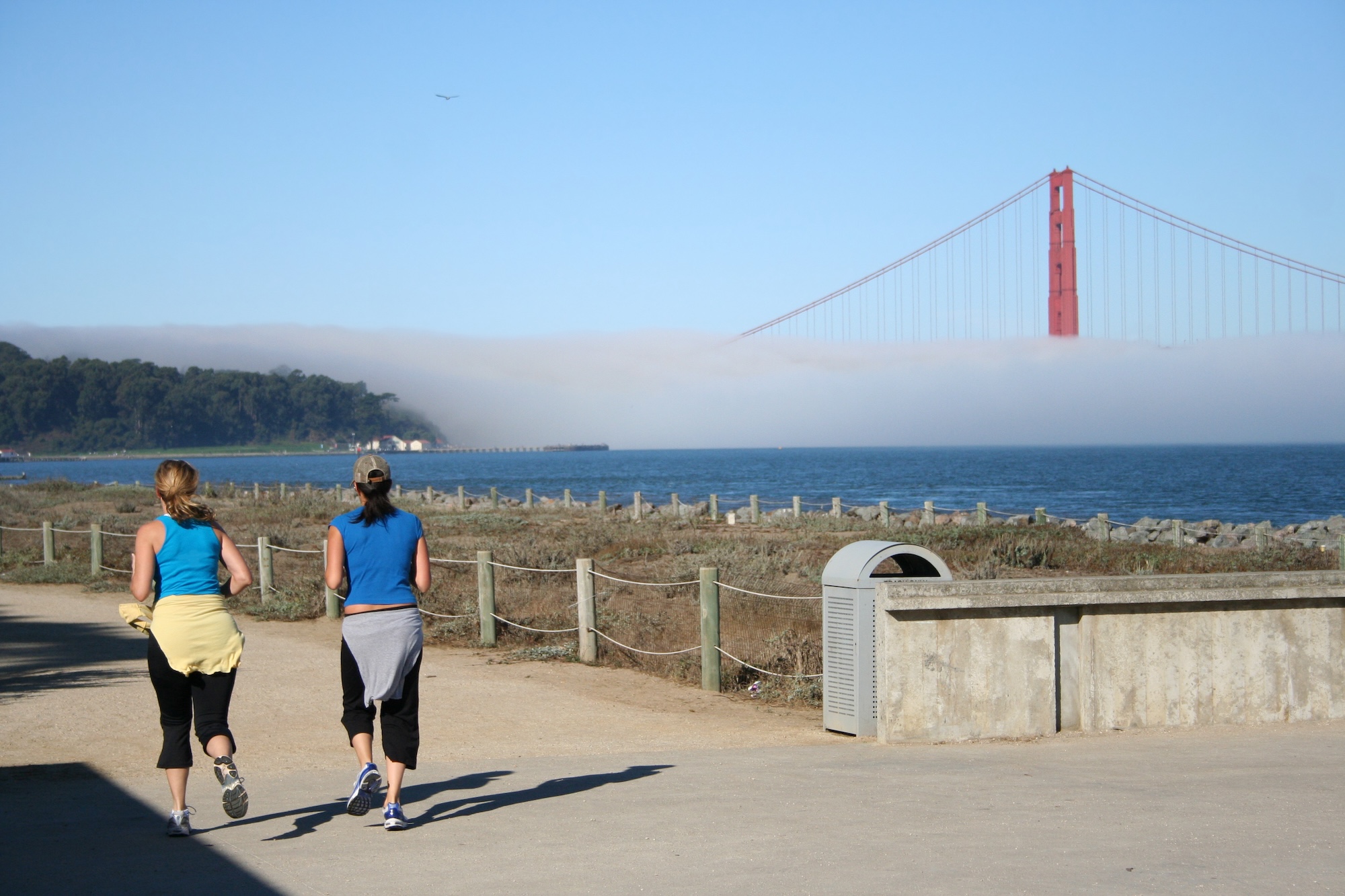 Two women jogging in San Francisco towards a fog-covered Golden Gate Bridge.