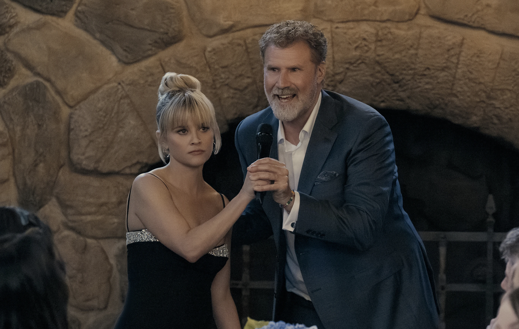 An unhappy looking blonde woman in a strapless evening dress holds onto a microphone as a man in a suit excitedly talks into it.