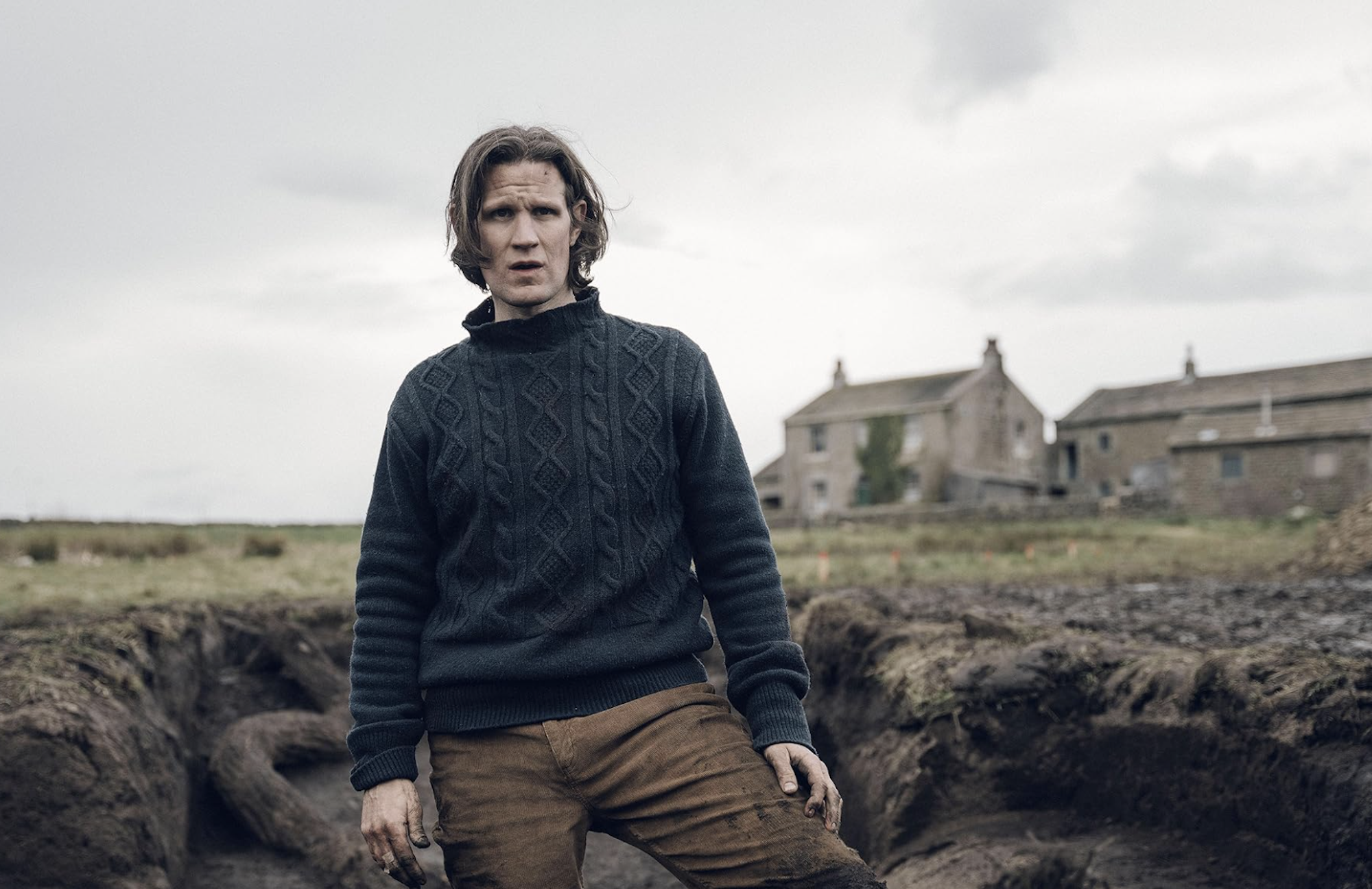 A man wearing brown cords and a sweater stands in a cold rural environment, with a big stone house behind him.