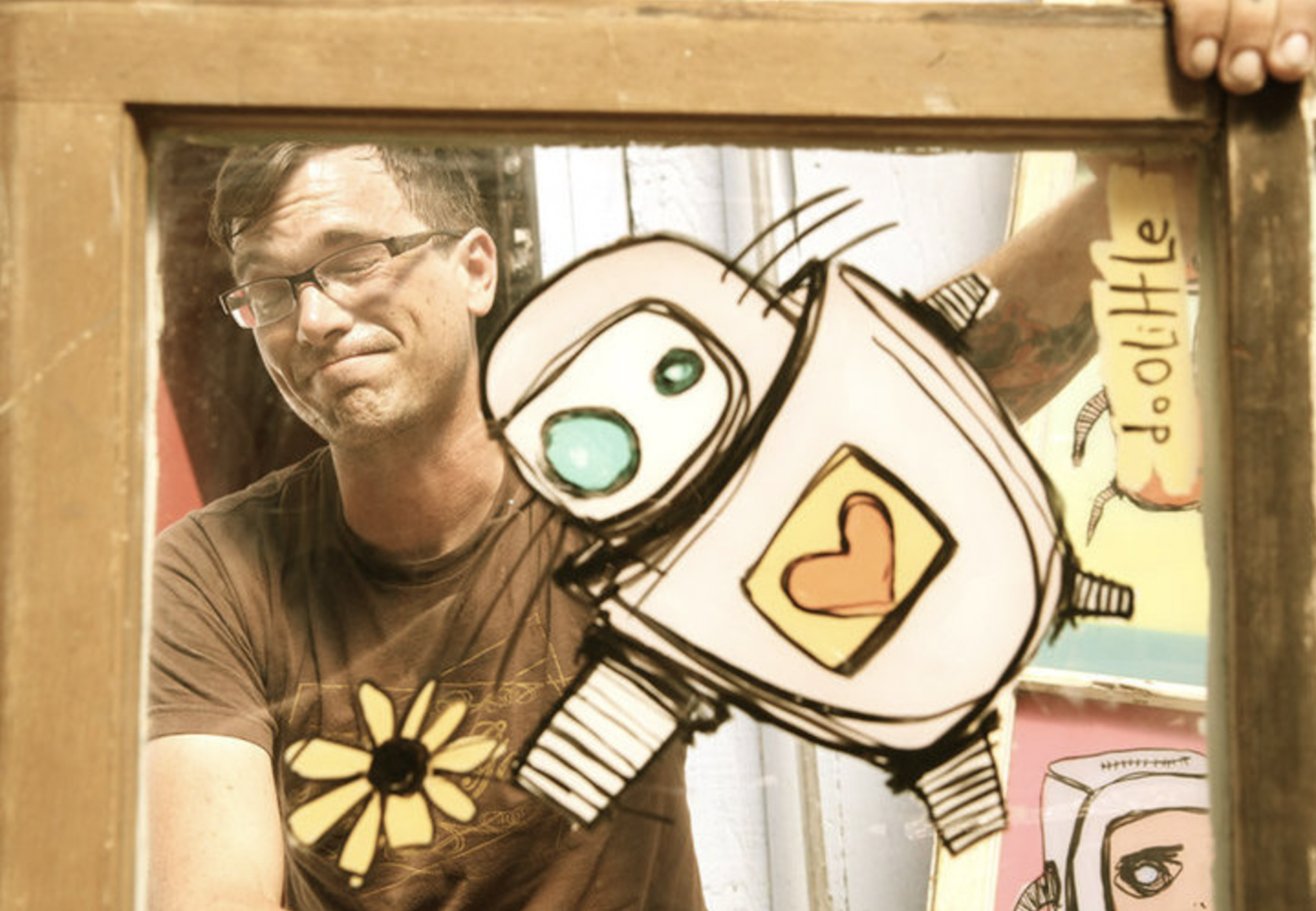 white man in glasses smiles while holding up window pane with painting of robot
