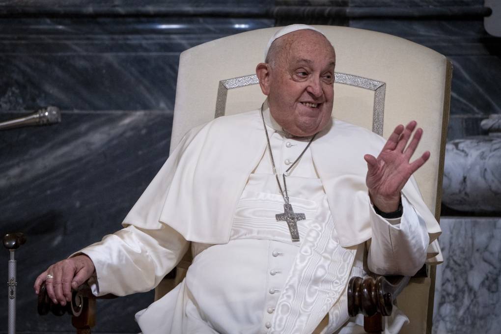The pope in white robes wearing a large crucifix smiles and gestures while sitting in a white chair.