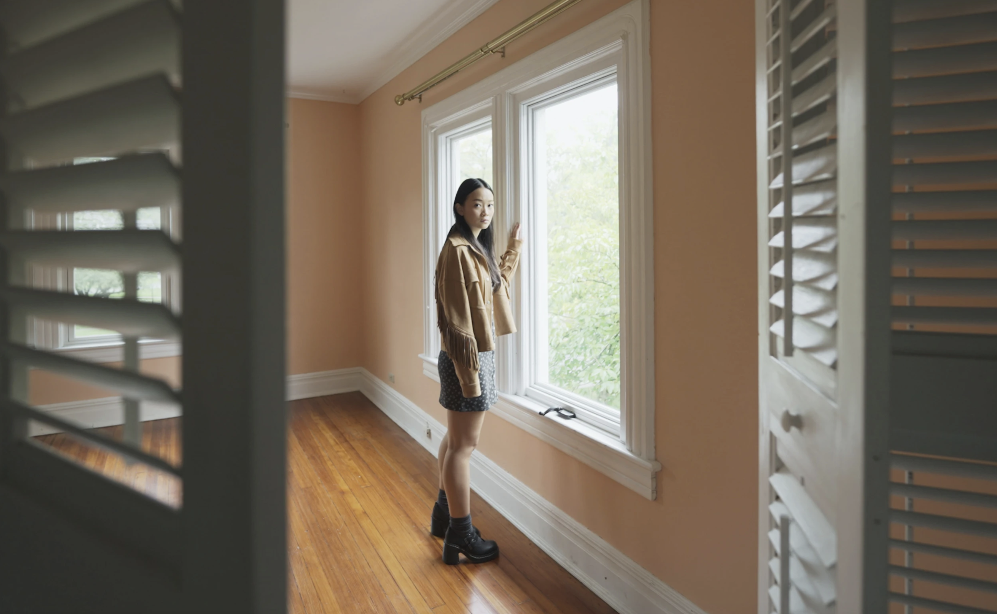 A young Asian woman stands before a window and gazes concerned into an open closet.
