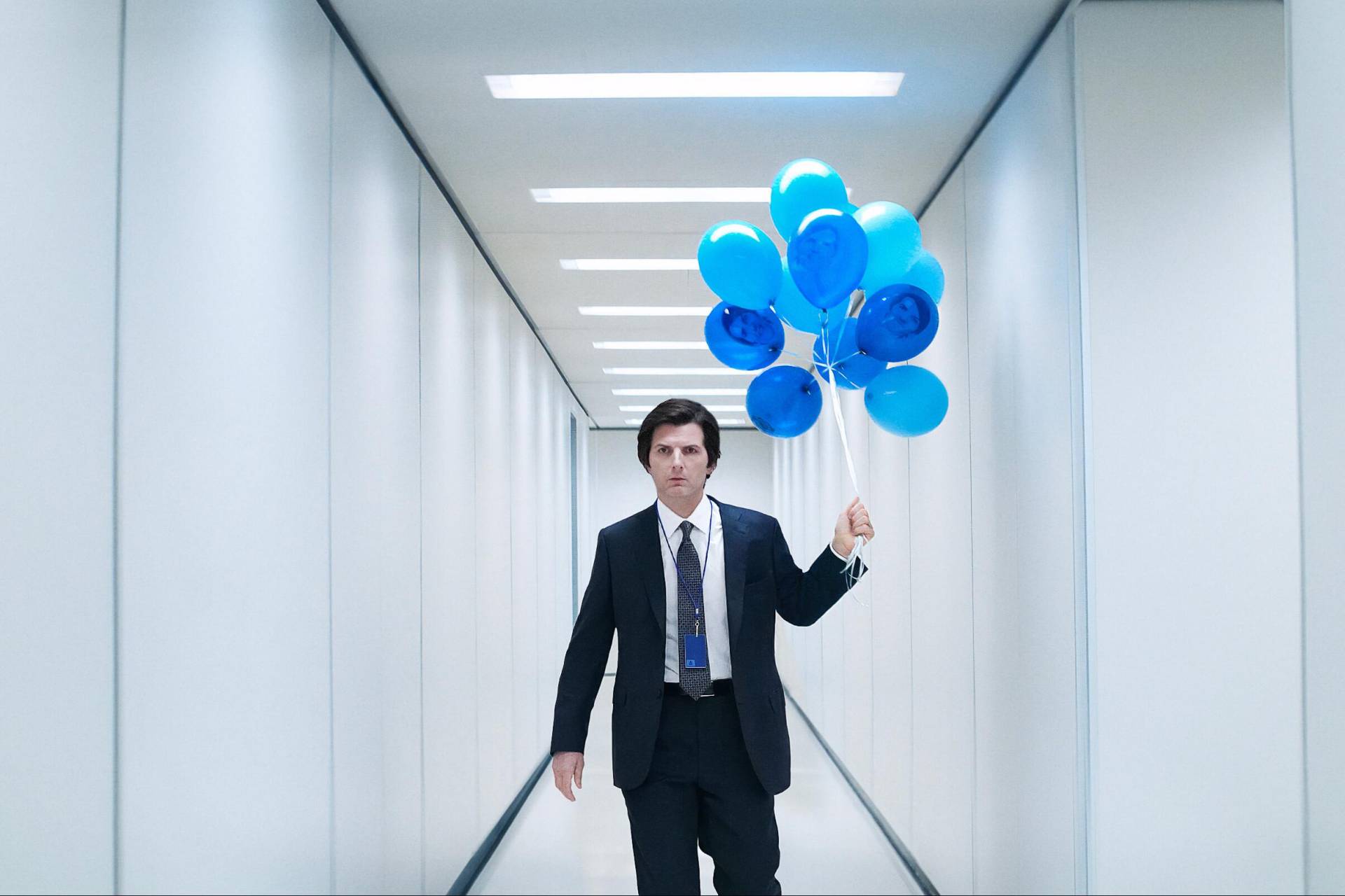 A white man in a black suit and blue tie walks down a stark white office hallway. He is carrying a bouquet of blue balloons in his left hand.