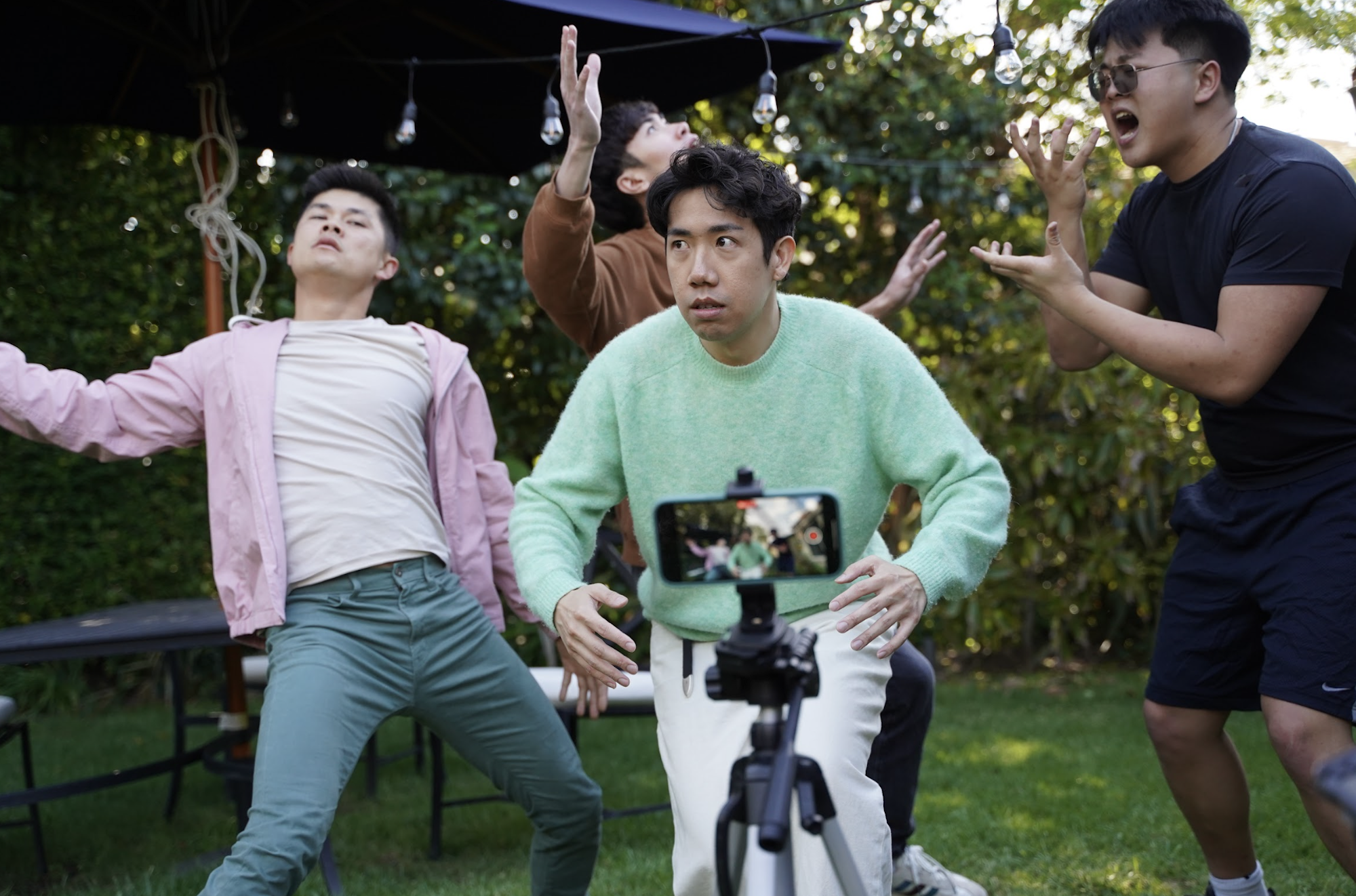 Four young Asian men gesture wildly as a camera phone on a tripod films them in a garden.