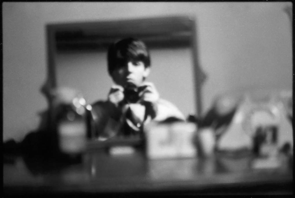 A young man reflected in a mirror, with a straight look on his face, somewhat out of focus, holding a camera.