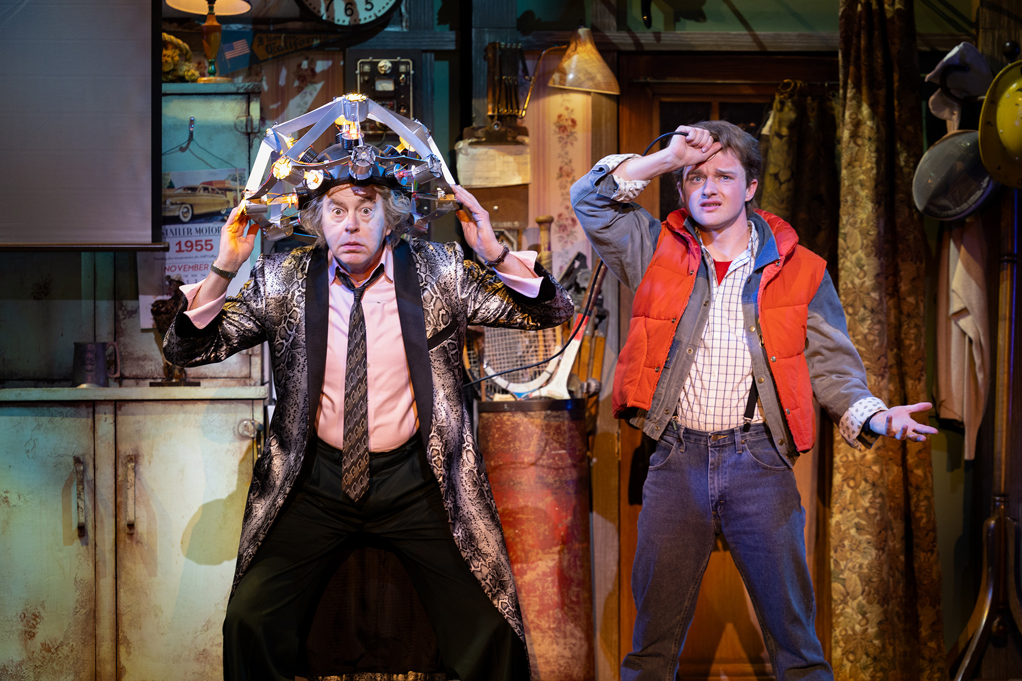 An older man with a scientific steel contraption on his head stands next to a young teenager in an orange vest and jeans, both with confused looks on their faces