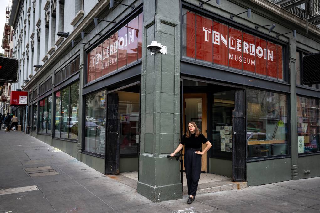 white woman in black stands against pillar of corner building underneath 'Tenderloin Museum' sign