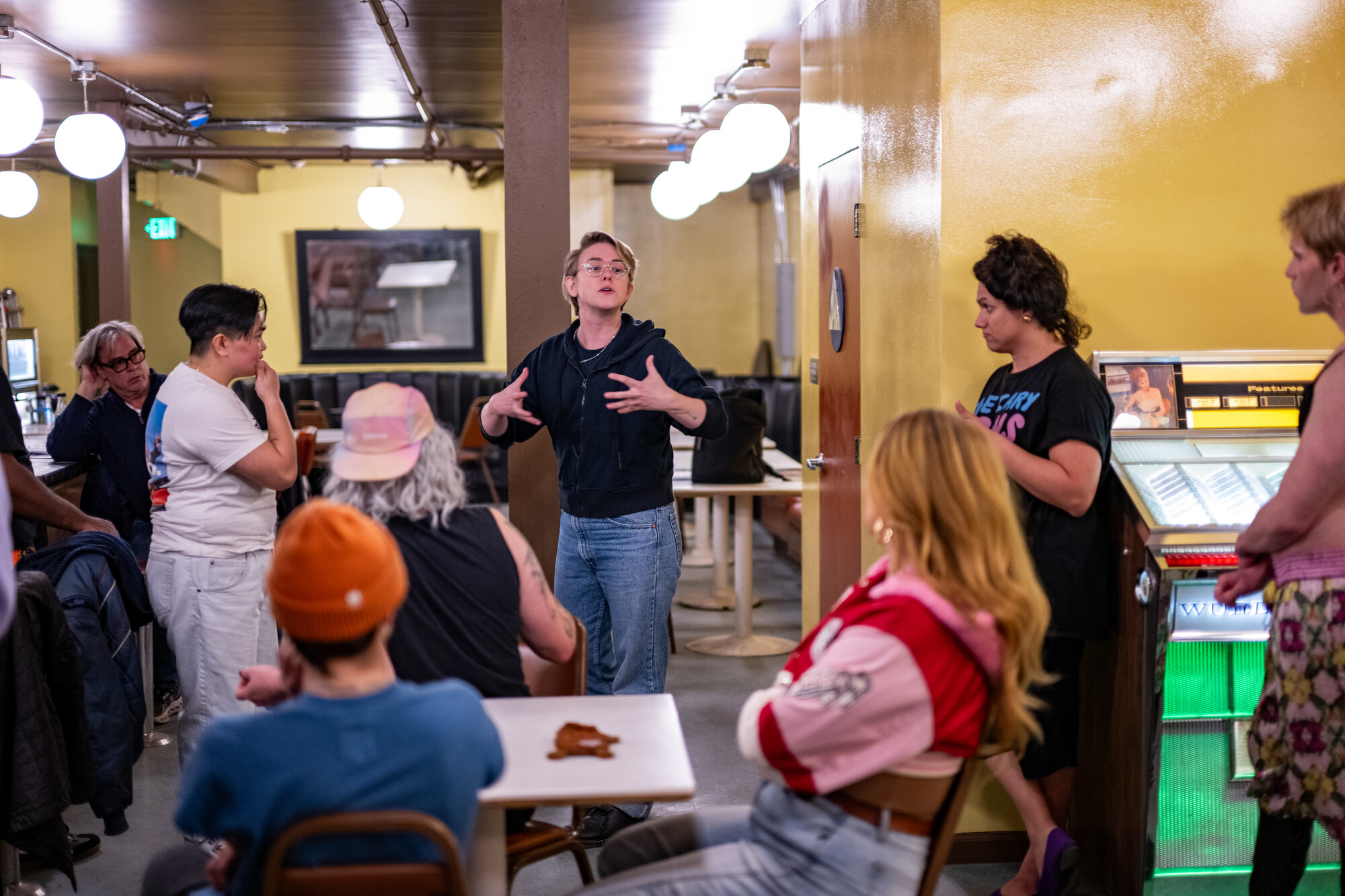 people look at person speaking, holding hands in emphasis, in a table-filled space