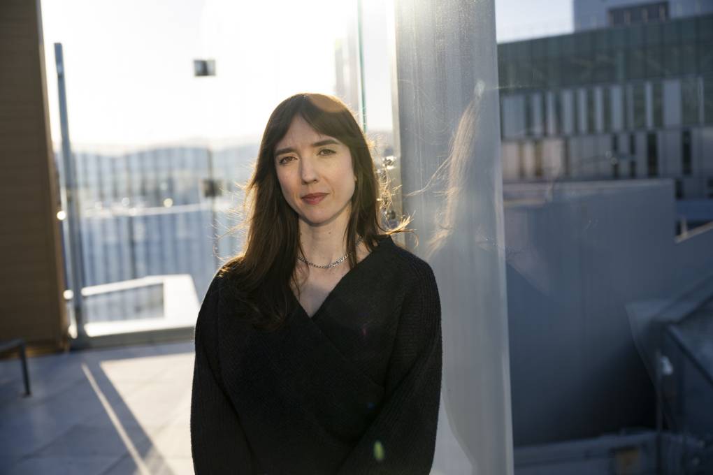 A woman in a black top looks into the camera as sun streaks in from an airport window