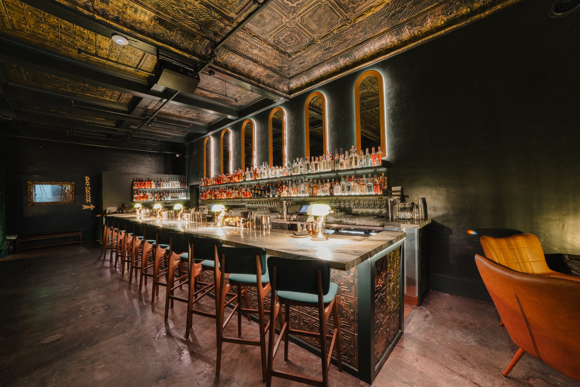 A view of a dimly lit bar with crown molding on the ceiling, velvet chairs and mid-century design accents.