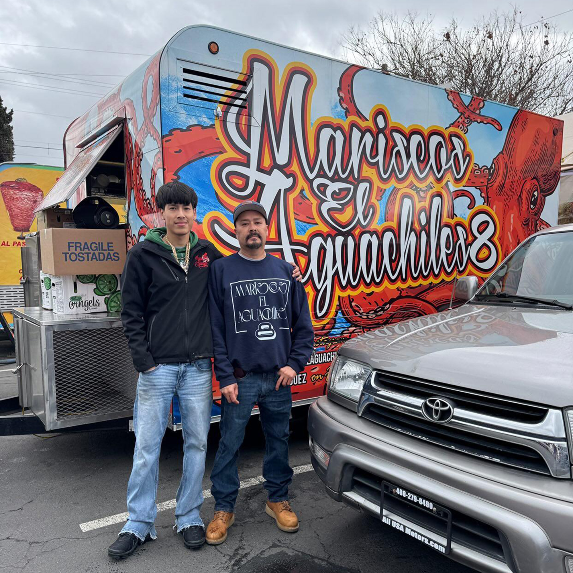 Two men pose for a portrait in front of their food truck. The painted lettering on the truck reads, "Mariscos El Aguachiles 8."