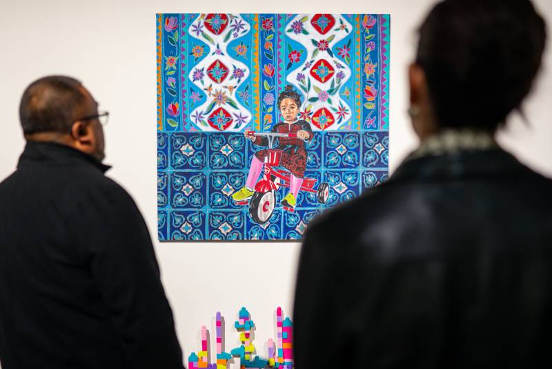 People look at an acrylic painting of a Palestinian toddler on a tricycle.