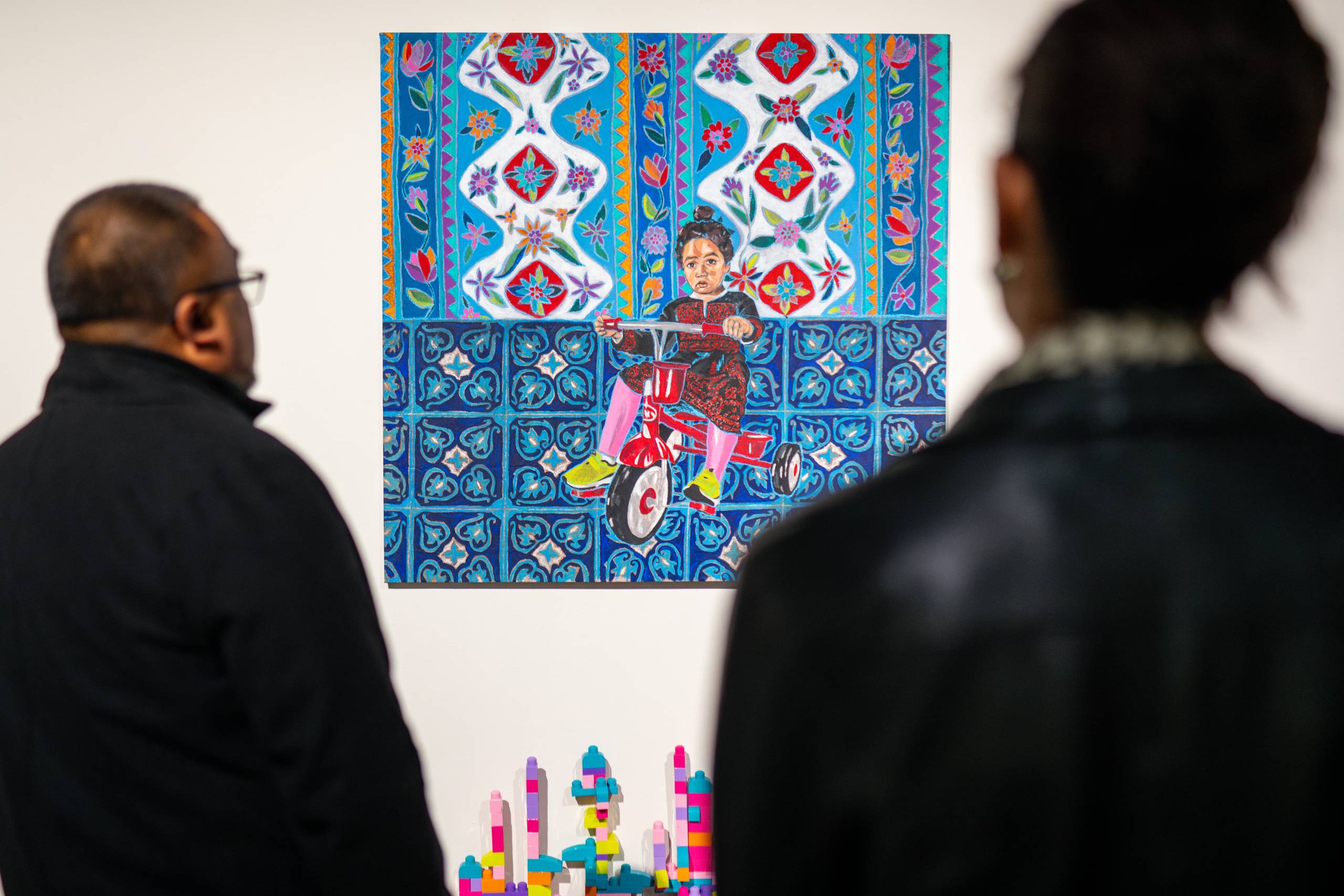 People look at an acrylic painting of a Palestinian toddler on a tricycle.