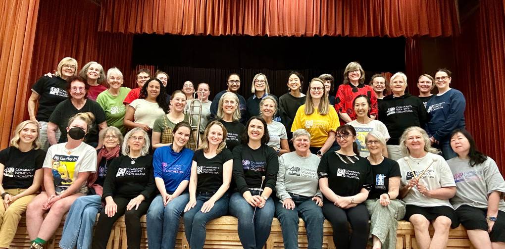 Women in casual T-shirts and jeans, some holding musical instruments, pose for a group photo.