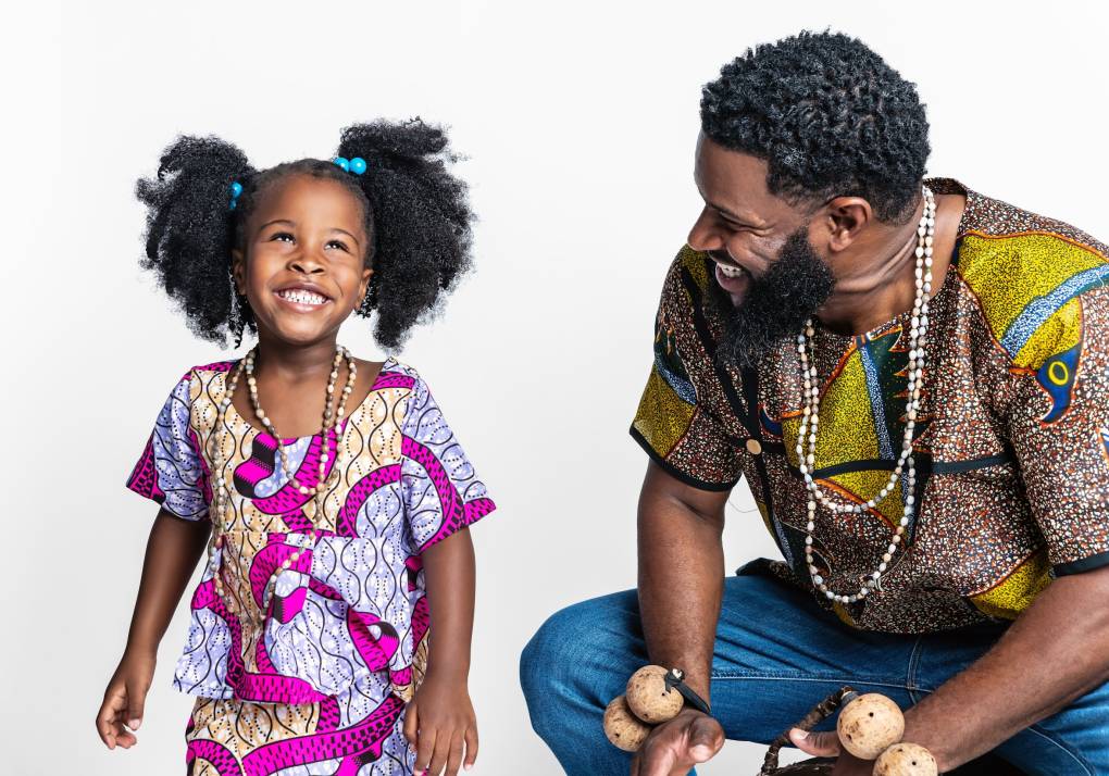 A girl smiles skyward as she stands next to her father, who is squatting and smiling while holding a drum.