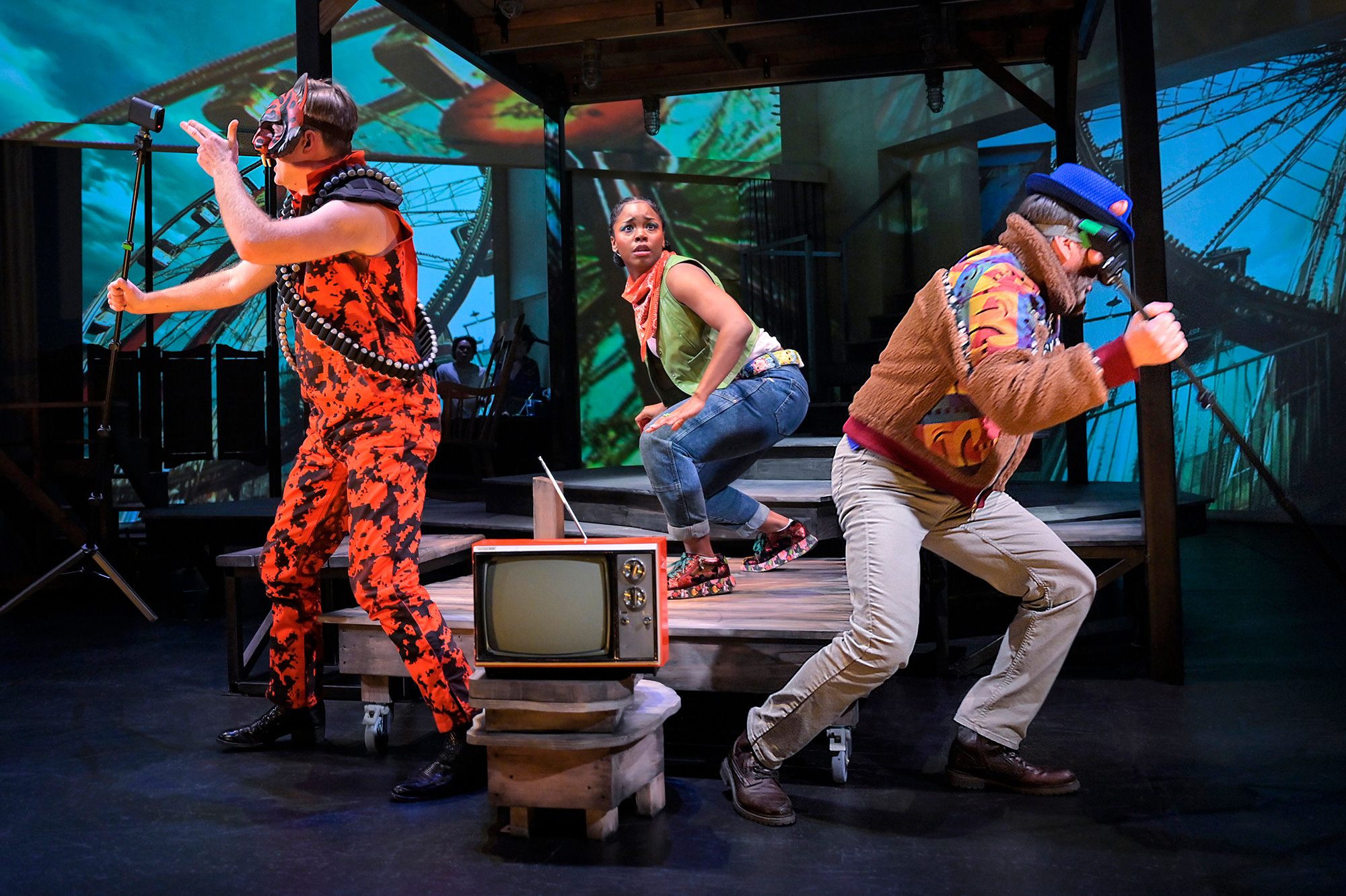 A young Black woman stands looking frightened at the center while two men dressed as video game characters run in opposing directions; lights and effects project upon a background screen