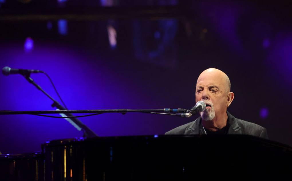 A bald man at a piano sings into a mic