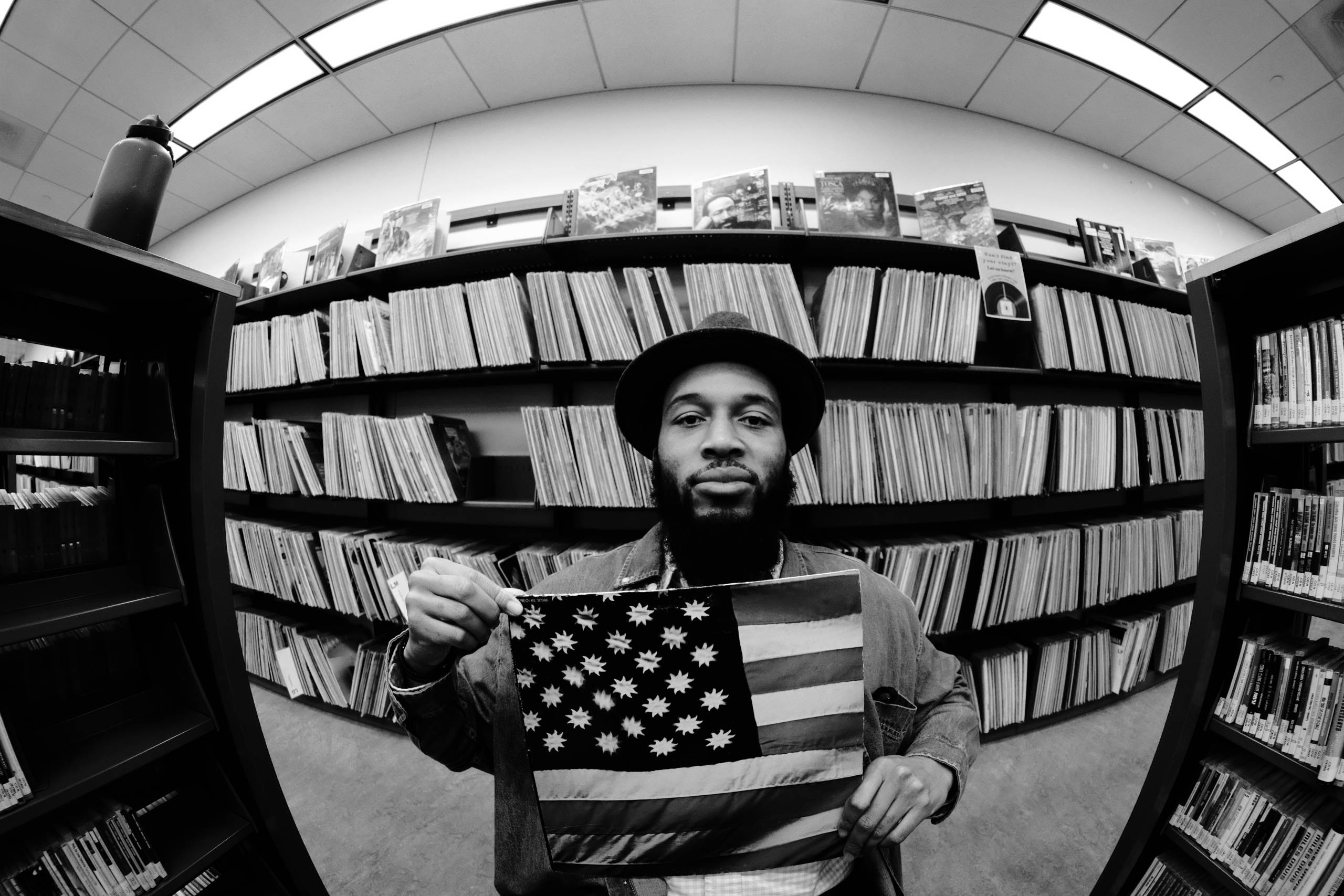 A black and white photo of a man holding a record while sitting in a room with a bunch of other records on the wall.