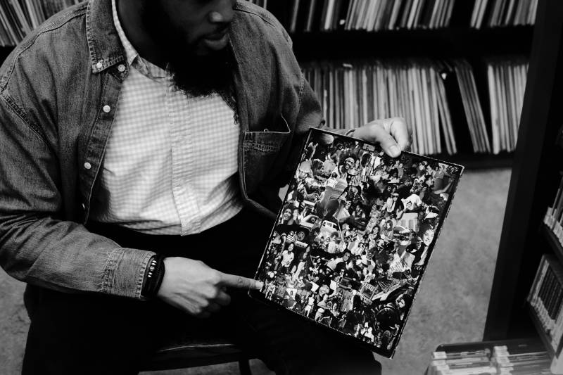 A black and white photo of a man holding a vinyl record, pointing to the image on the album's cover. 