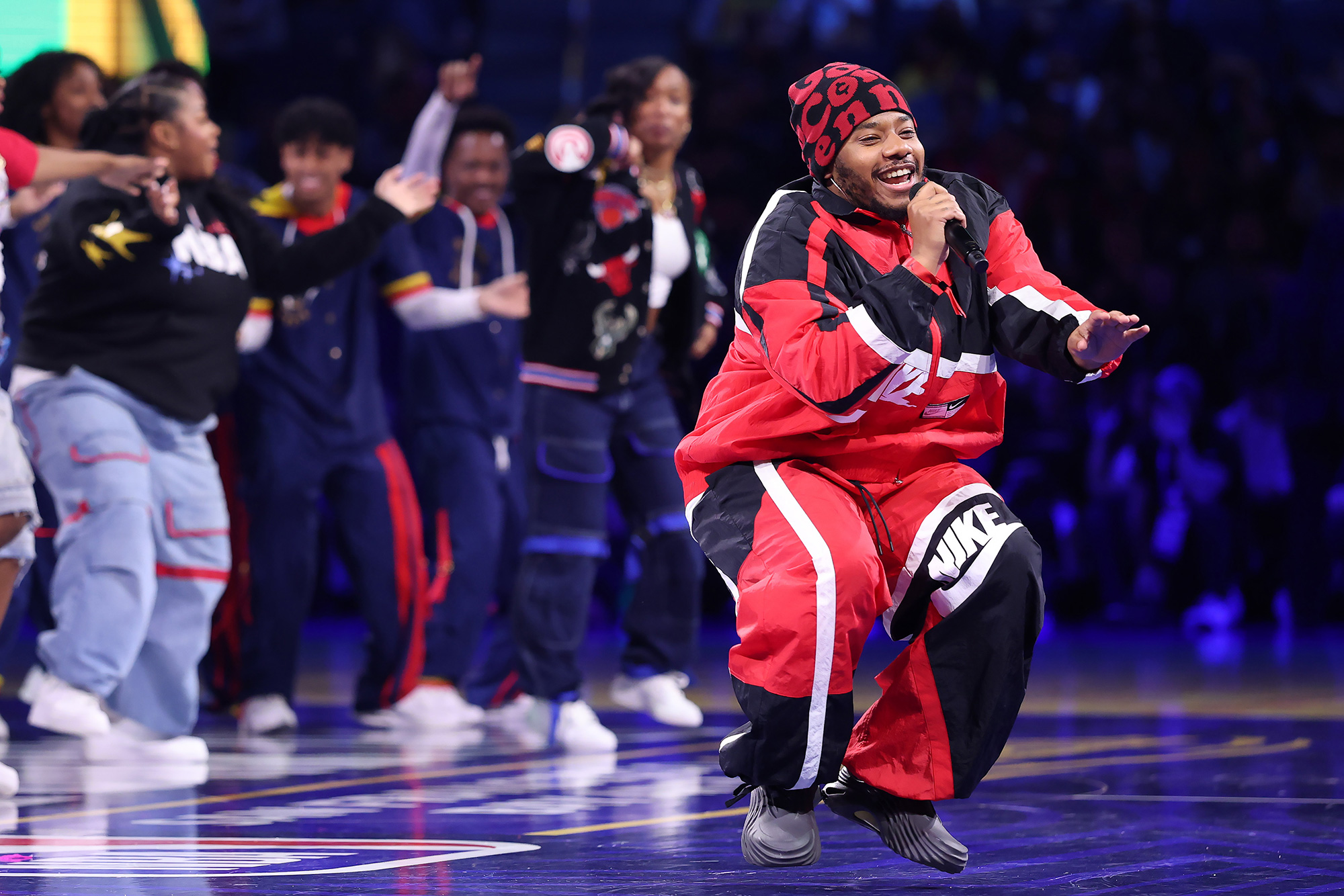 A young Black man in an oversized athletic suit branded with Nike logos smiles and raps into a microphone on a basketball court, with dancers in the background
