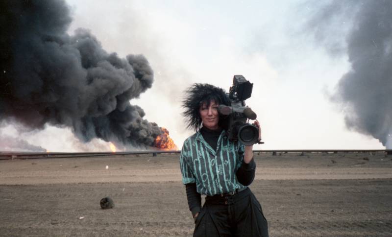 woman with wild black hair stands with camera, smoke from fires in background