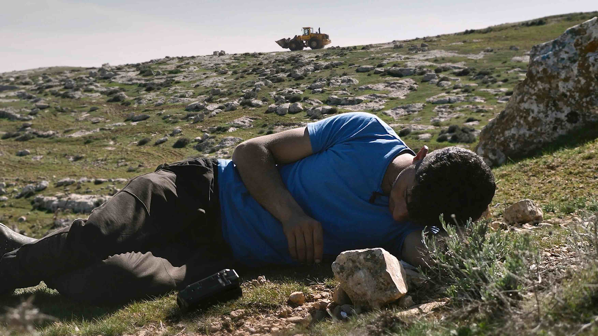 man in blue shirt lies on rocky ground, bulldozer on hilltop behind him